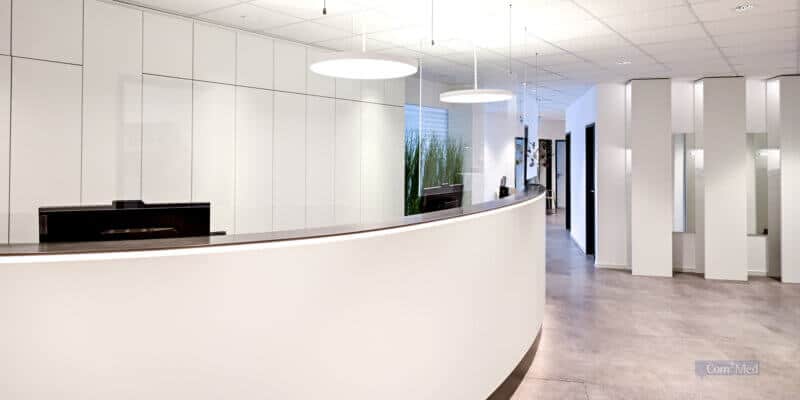 Geräumiger Empfangsbereich mit geschwungenem weißen Tresen, spiegelnden weißen Wandschränken, rundum verteilten Deckenleuchten und einer Glasfront mit Blick auf Grünpflanzen.
