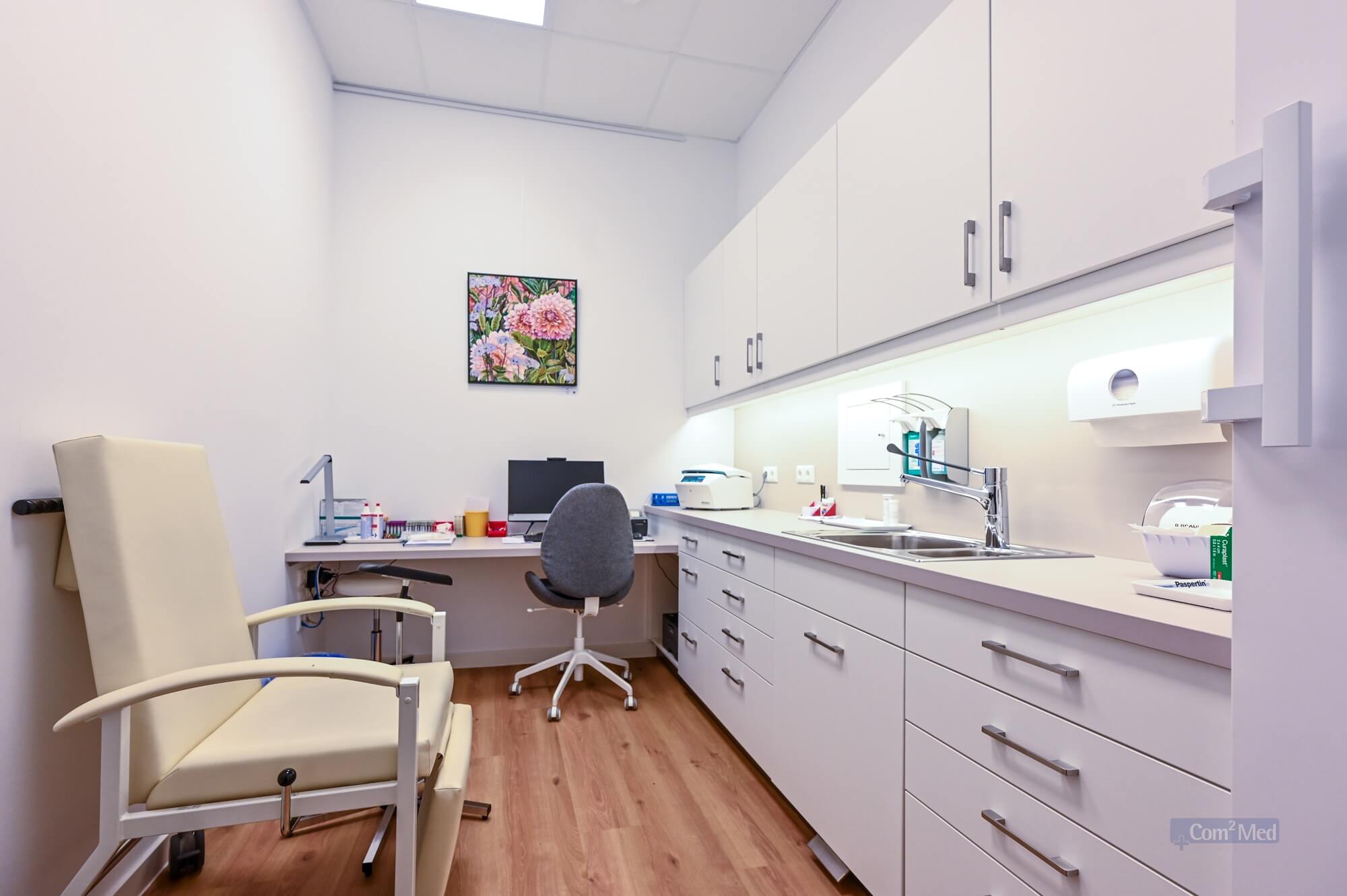 Behandlungsraum mit cremefarbenem Untersuchungsstuhl, Schreibtisch mit Computer und medizinischem Equipment, weißer Einbauschrank mit Spüle und aufgehängtem Blumenbild an der Wand.