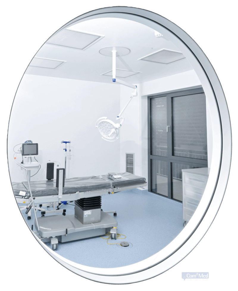 Blick durch ein rundes Fenster auf einen leeren Praxiszimmer mit einem OP-Tisch, einer Operationslampe und medizinischer Ausrüstung in einem hellen, klinisch weißen Raum.