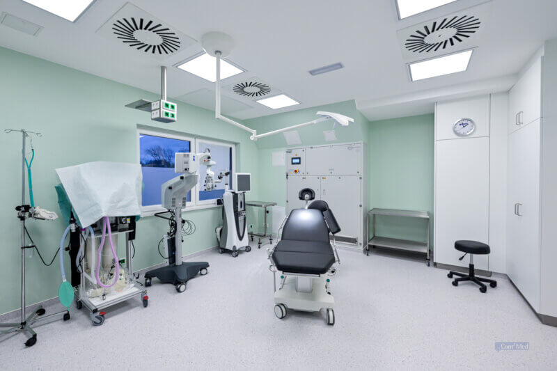 Modernes Behandlungszimmer mit OP-Tisch in der Mitte, umgeben von medizinischen Geräten, Überwachungsmonitoren, Operationsleuchten und grünen Wänden, die eine beruhigende Atmosphäre schaffen.