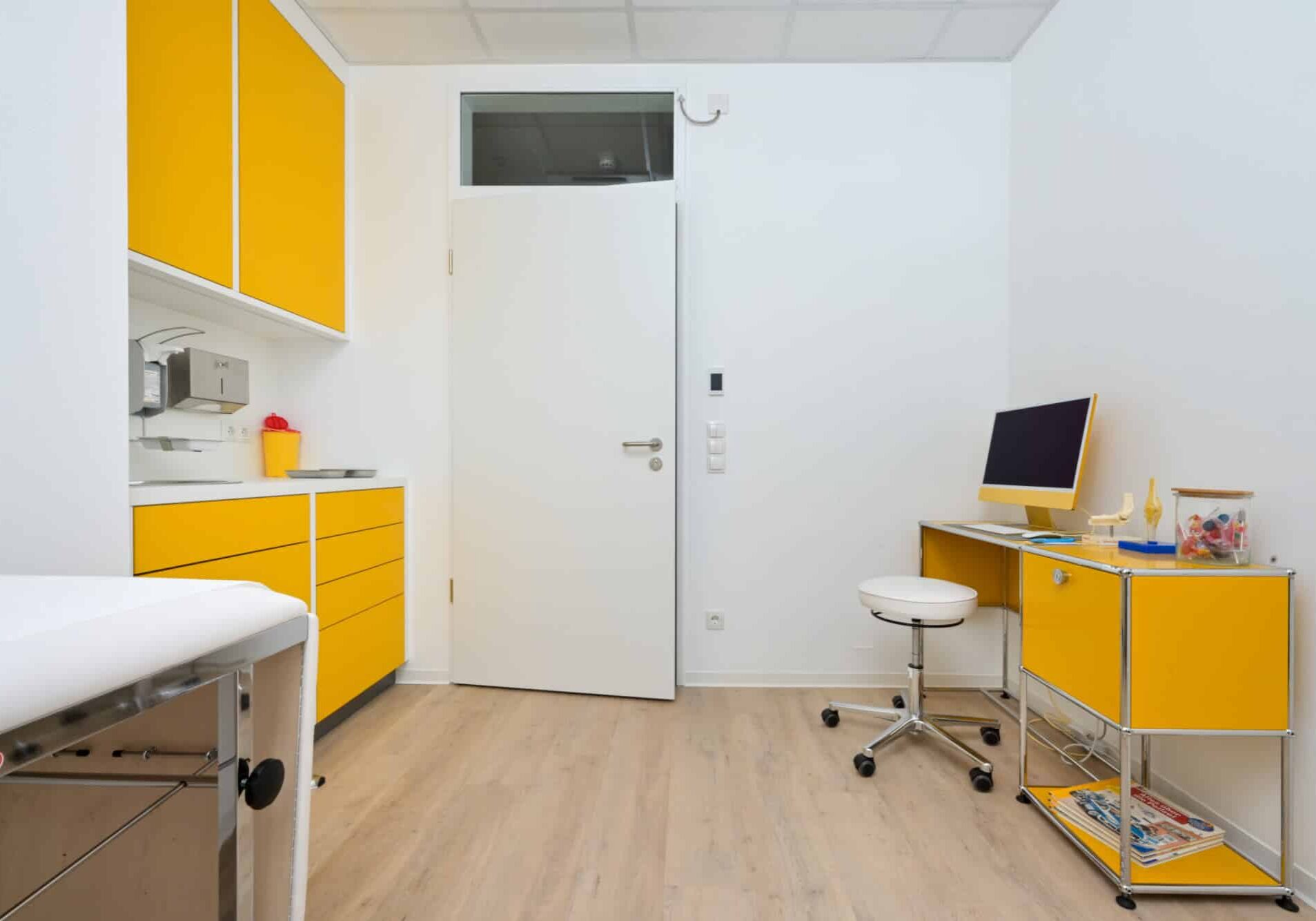 Behandlungsraum mit weißer Liege, gelben Schränken, weißem Hocker und Schreibtisch mit Computer und medizinischen Utensilien.