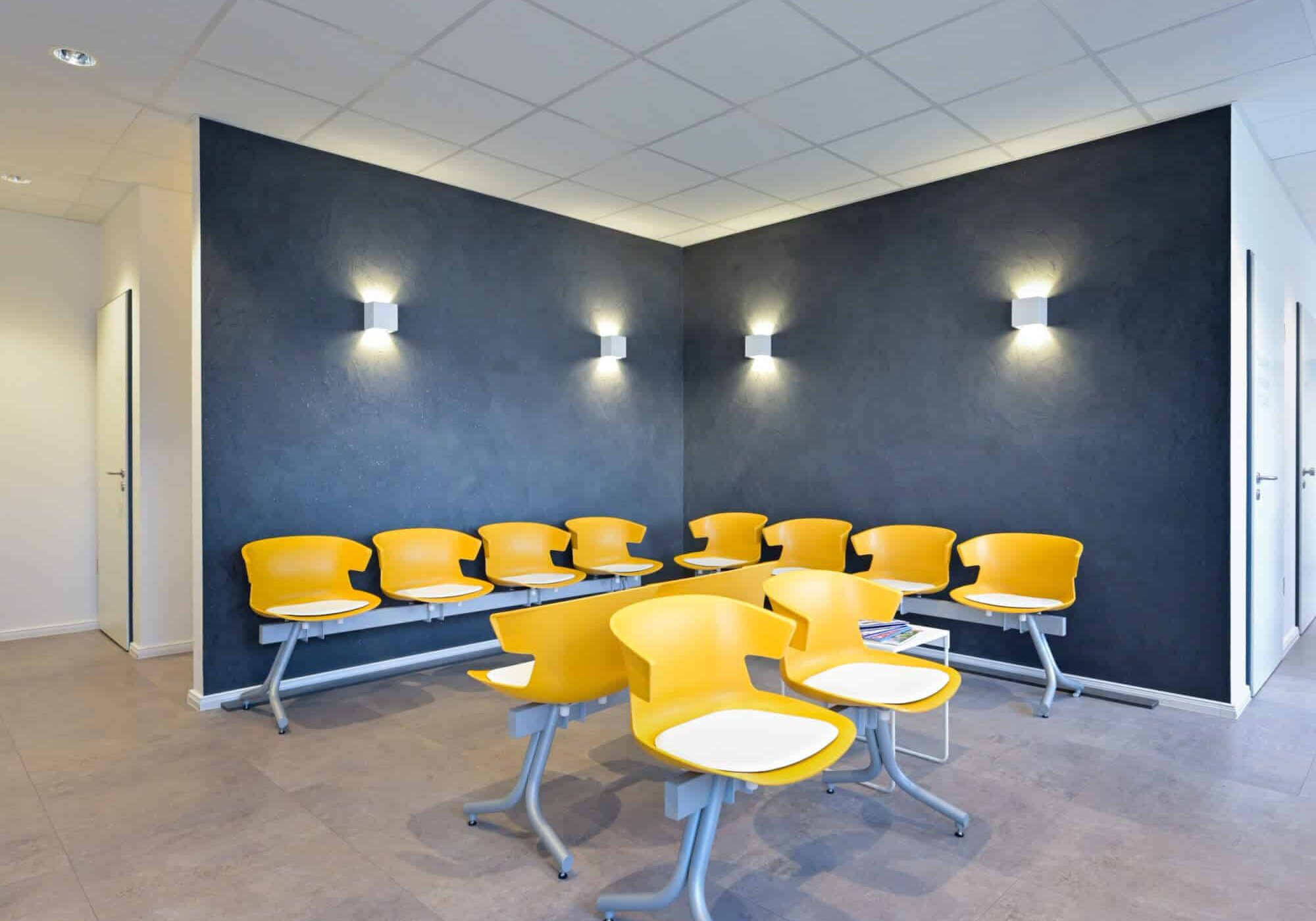 Wartezimmer mit gelben Stühlen an einem grauen Tisch, dunkelgrauer Akzentwand mit aufgesetzten Lampen und zwei Türen im Hintergrund.